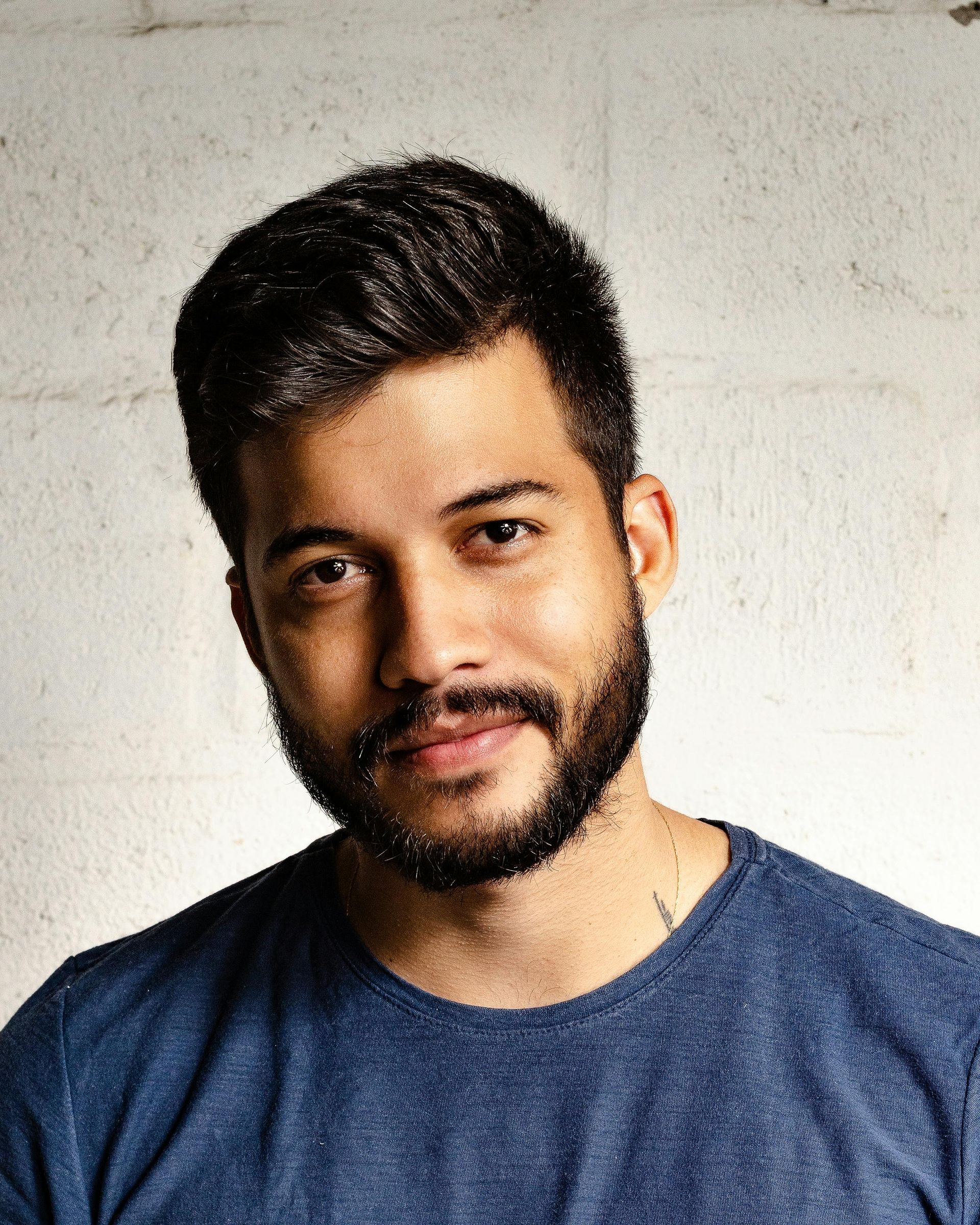 Man with dark hair and beard, wearing blue shirt, smiling against a textured white background.