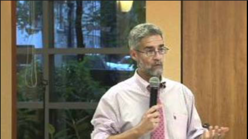 A man in a pink shirt and tie is speaking into a microphone.