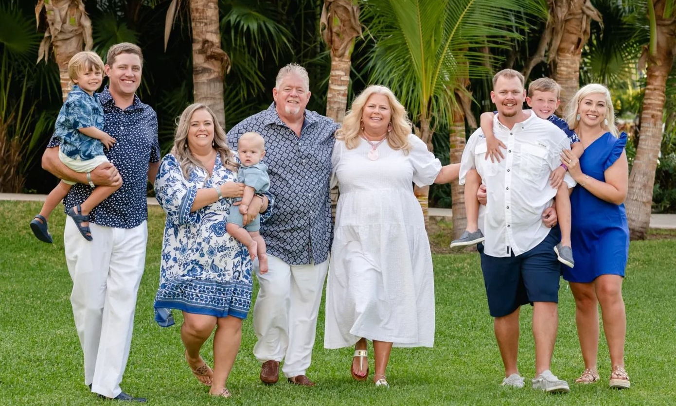 A family poses for a group photo on a grassy lawn with palm trees in the background, smiling together.