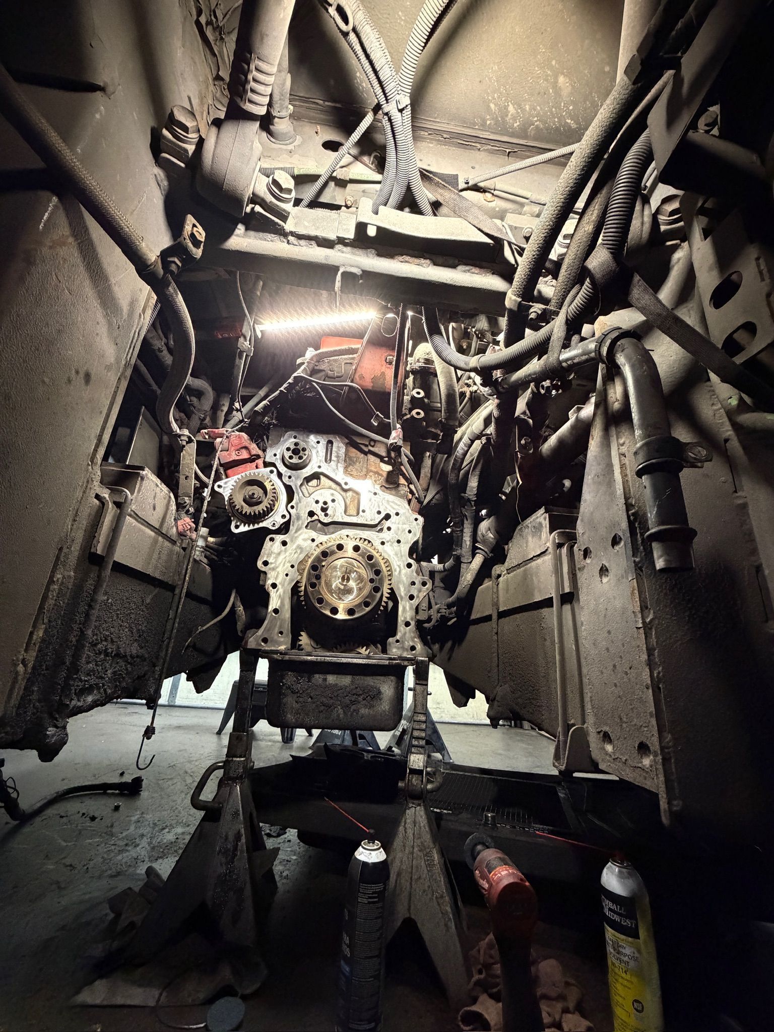Inside a cramped industrial tunnel, a machine engine is lit by a bright work light and surrounded by cables.