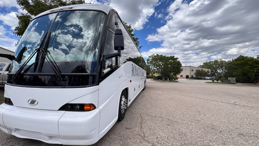 White coach bus parked on a gravel lot under a cloudy sky with trees and a building in the background