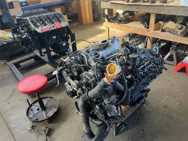 Two disassembled car engines on stands in a workshop, with tools and shelves in the background.