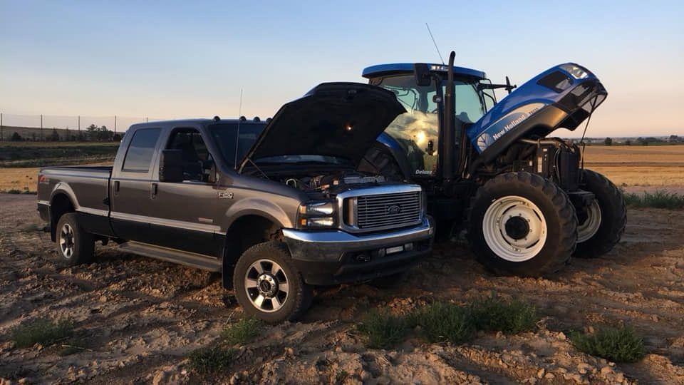 Pickup truck and backhoe collide on a dirt road at sunset, with the backhoe’s bucket raised over the truck’s hood.
