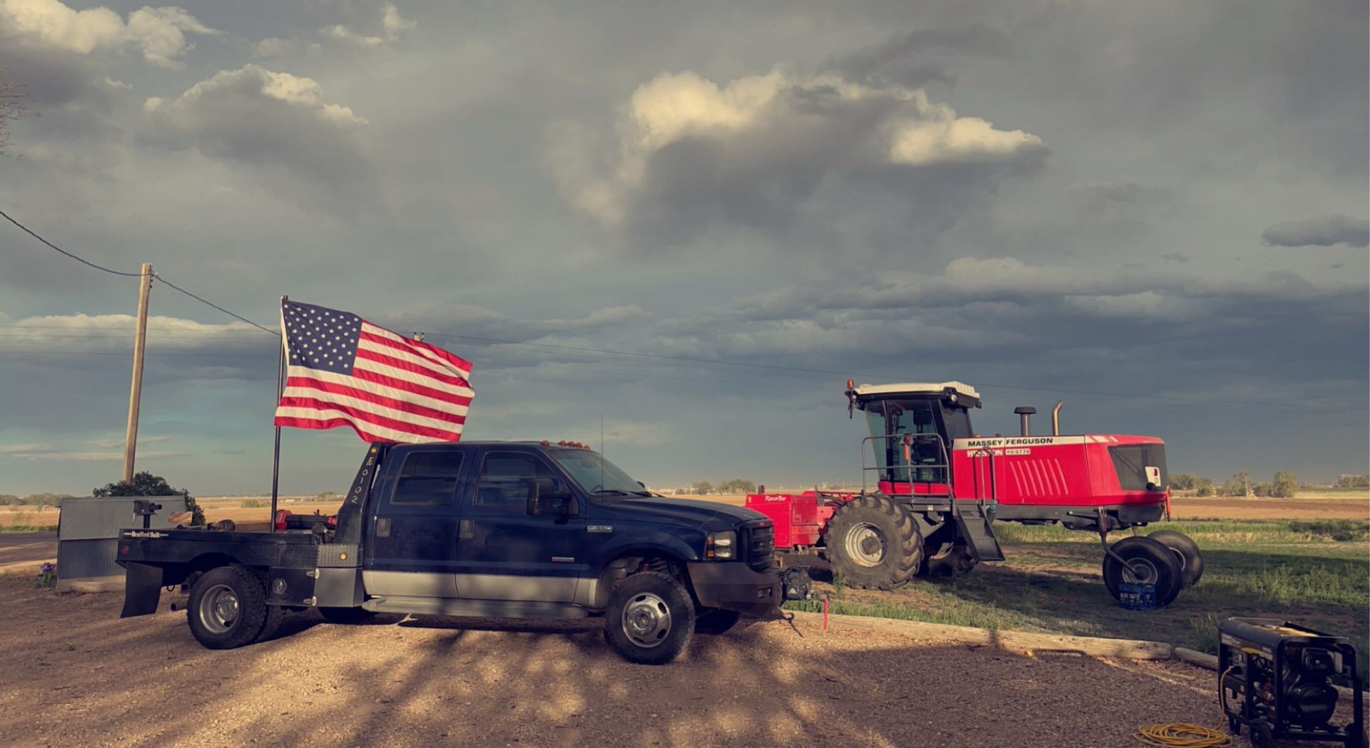 Pickup truck with large American flag beside a red combine in a muddy field under cloudy skies