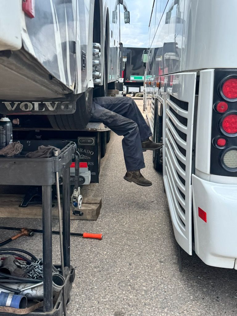 Person working under a white Volvo truck beside another vehicle in a parking lot.