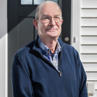 A man wearing glasses and a blue sweater is standing in front of a door.