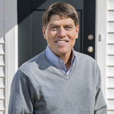 A man in a grey sweater is standing in front of a black door.