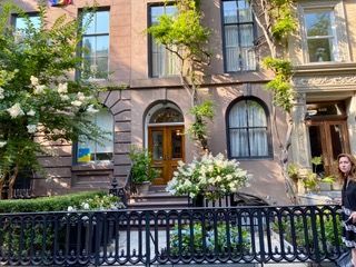 Brownstone building with arched doorway and black wrought iron fence. Woman stands on the right. Lush greenery.