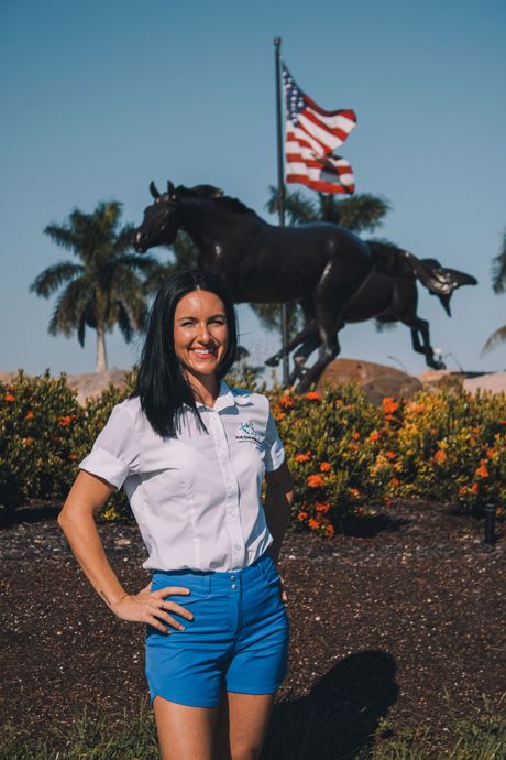 A woman in a white shirt and blue shorts is standing in front of a statue of a horse.