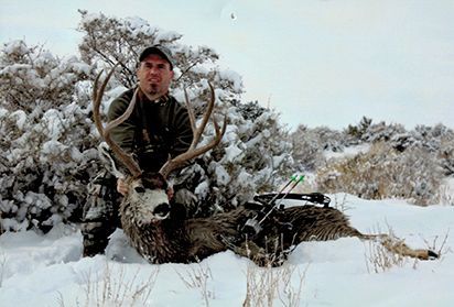 Hunter kneels in snow next to a deer with large antlers; bow and arrow nearby.