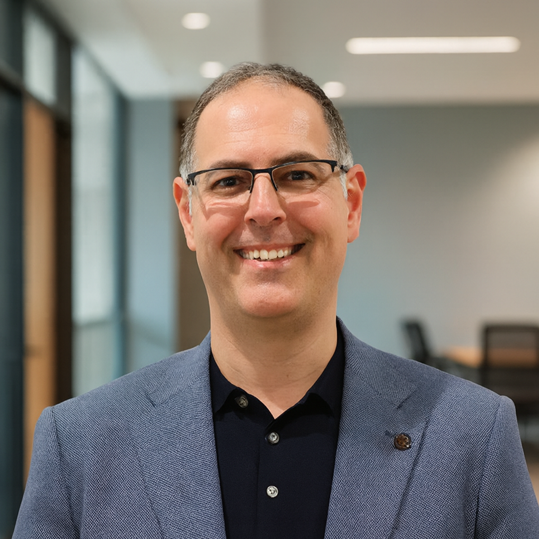 Man in glasses smiling, leaning against a brick wall, wearing a collared shirt.