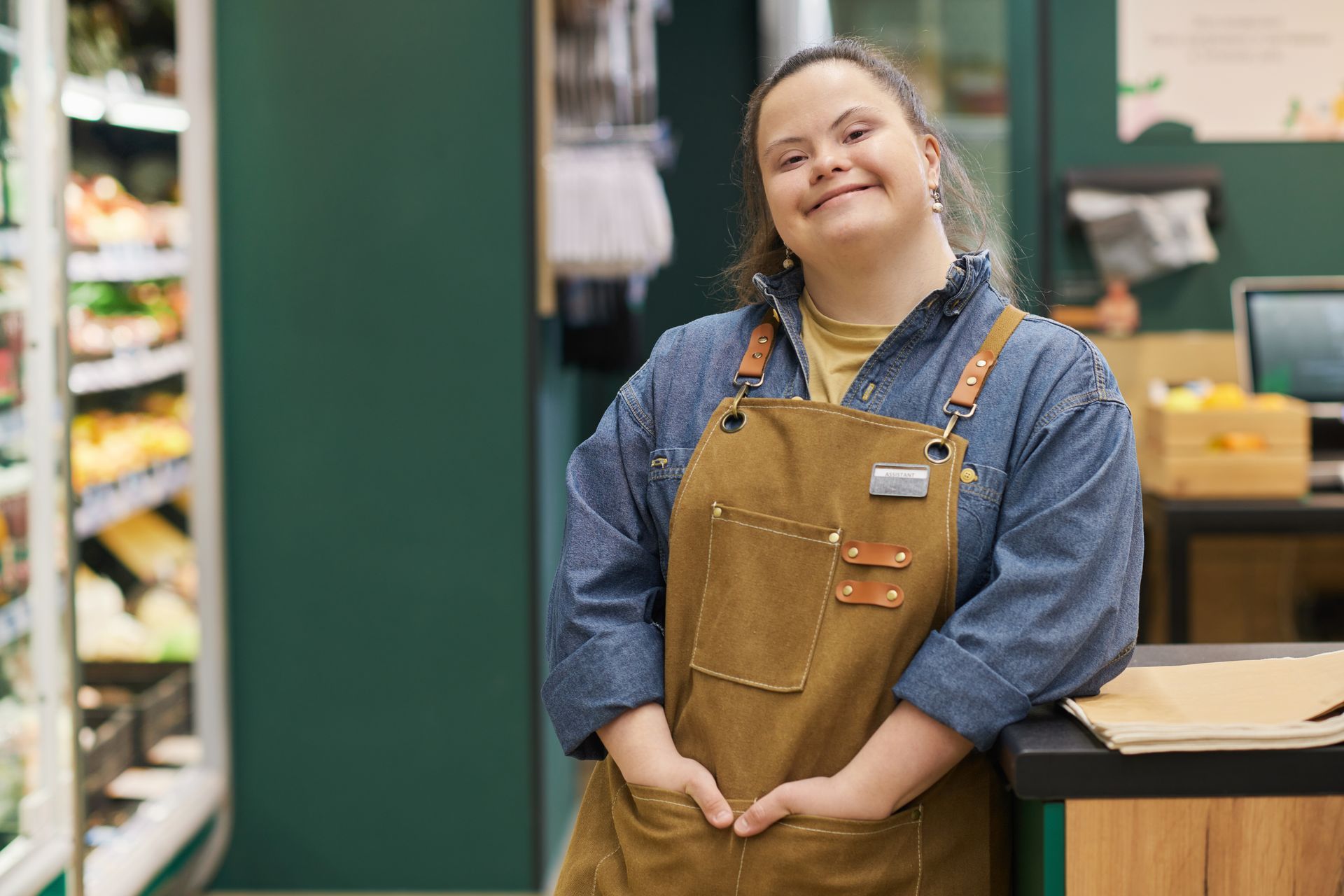 Woman with Down syndrome wearing an apron smiles at the camera in a grocery store setting.