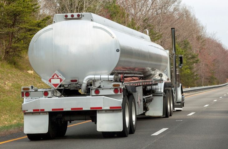 Tanker truck driving on a highway, carrying a cylindrical tank, red hazard label, trees in background.