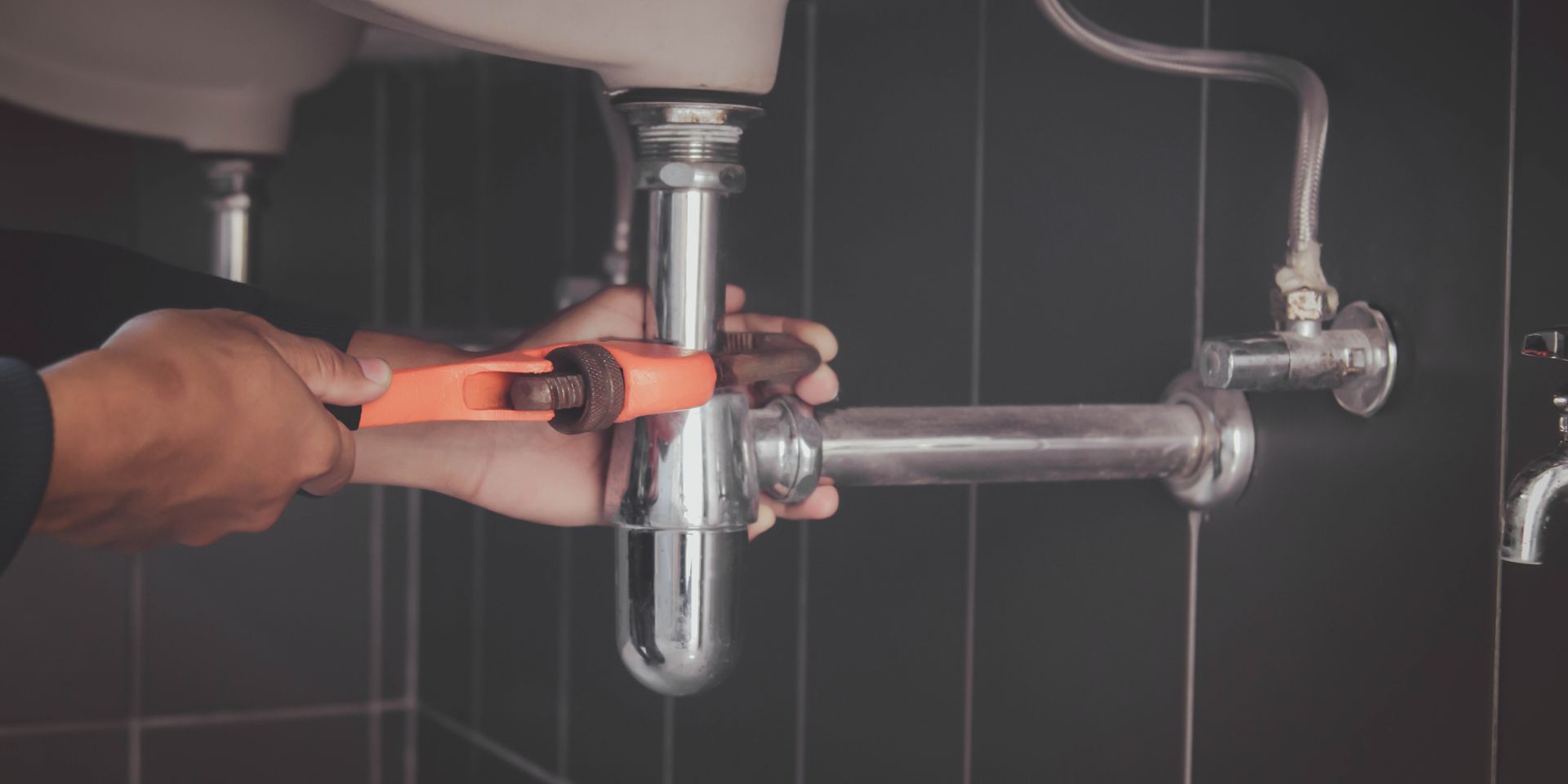 Hands using an orange wrench to tighten a pipe under a sink in a bathroom.