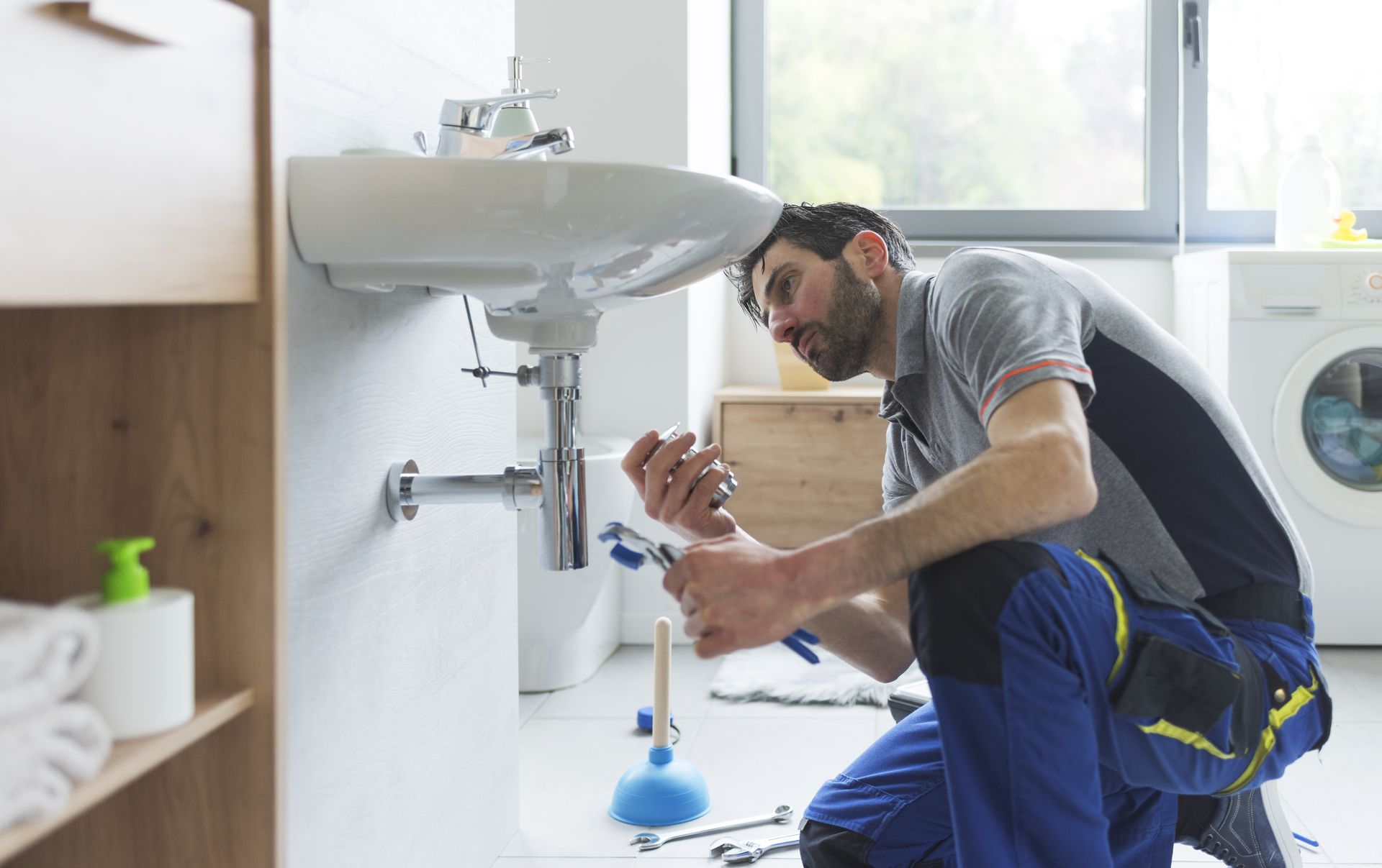 A plumber kneels, repairing a bathroom sink with tools. He wears blue pants and a gray shirt.