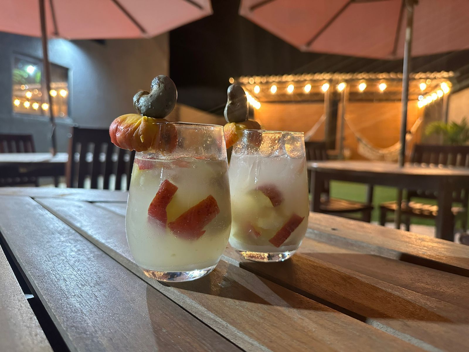 Dois coquetéis decorados com frutas e nozes sobre uma mesa de madeira ao ar livre à noite.