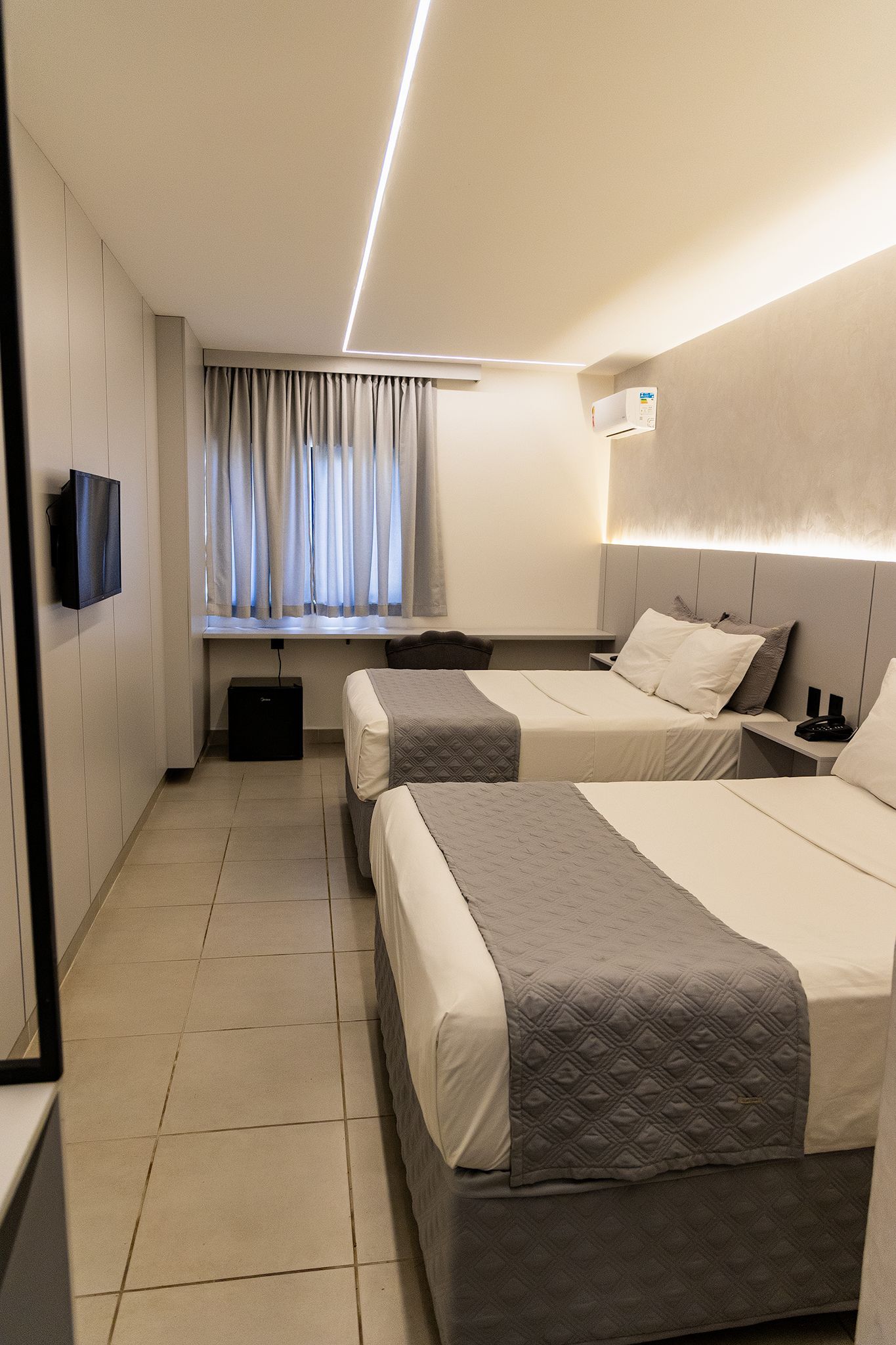 Dois quartos de hotel com decoração em tons de cinza e branco e uma TV fixada na parede.