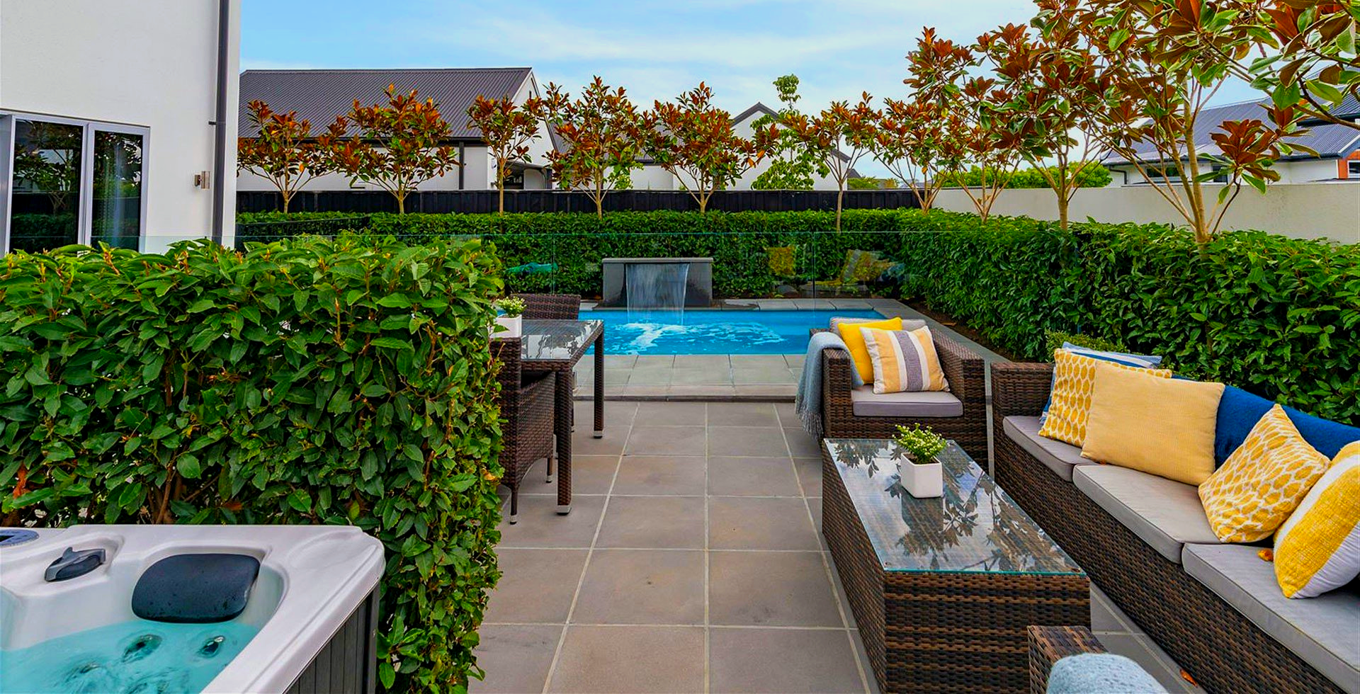 Patio with hot tub, pool, and lounge seating surrounded by greenery and trees.