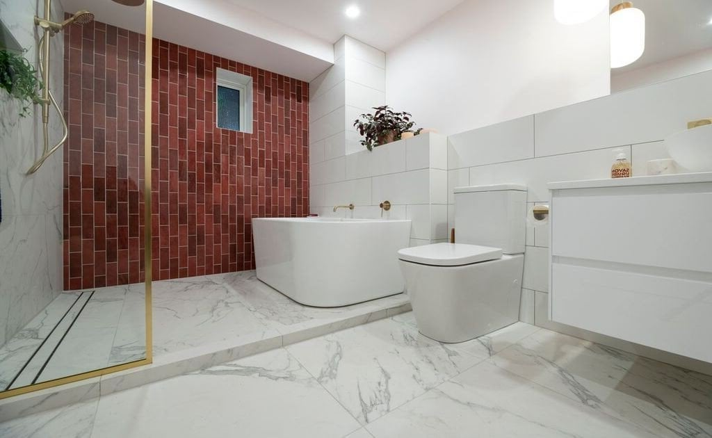 Modern bathroom with a white tub, toilet, and vanity against a red brick accent wall.