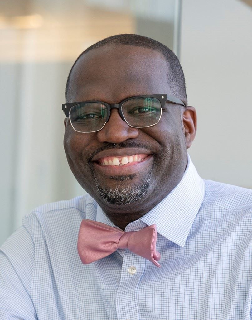 Smiling man in glasses and pink bow tie in a light blue shirt, seated indoors.