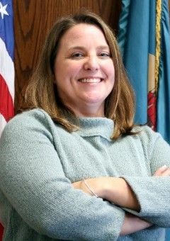 Smiling woman in a light blue sweater standing with arms crossed in front of flags