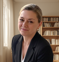 Portrait of a person in a dark blazer, smiling in a bright room with bookshelves behind them