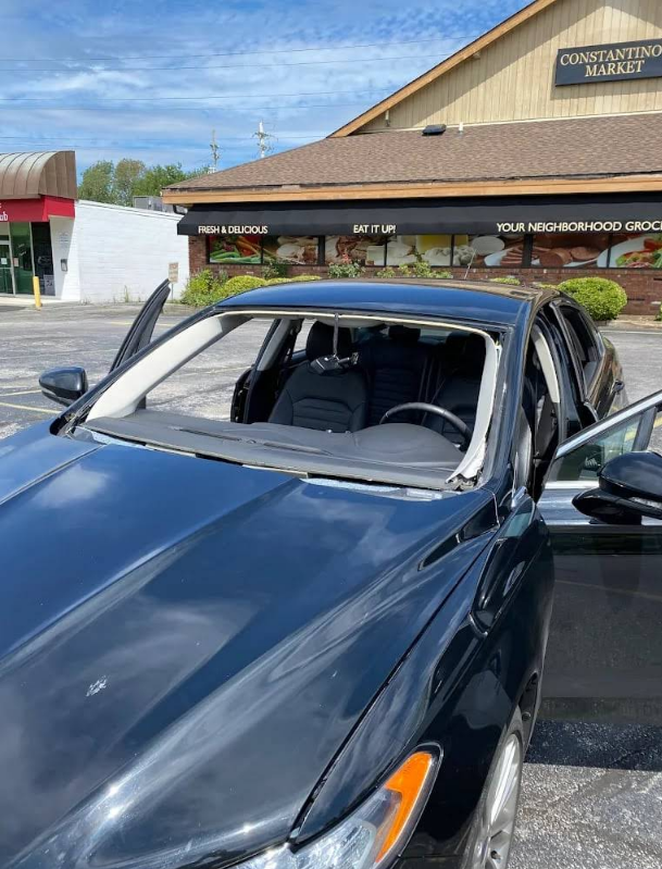 Black car with shattered windshield, doors open, parked in front of a market.