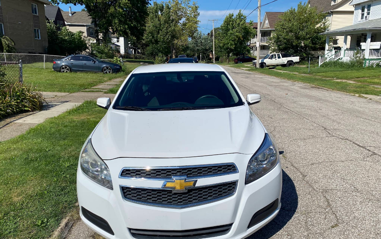 White Chevrolet Malibu parked on a residential street; houses and trees in the background on a sunny day.