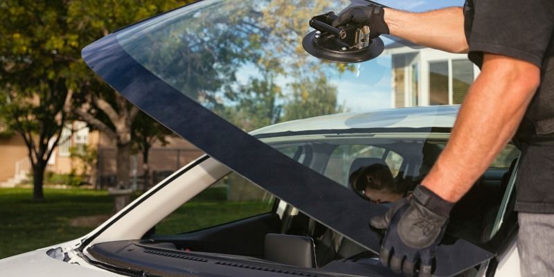 A person with black gloves installing a windshield on a white car outdoors.