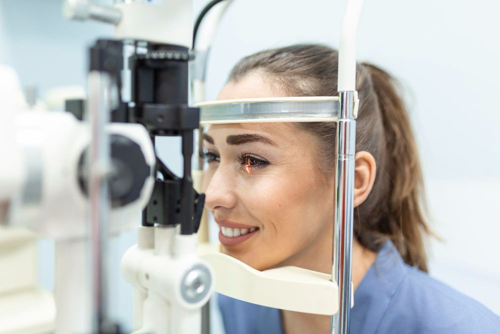 Woman at an eye exam looking into a slit lamp, smiling in a bright clinic.