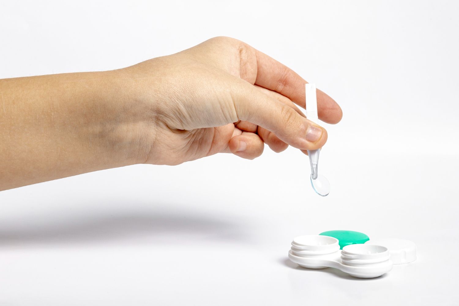 Hand placing a contact lens over an open lens case with green liquid on a white background