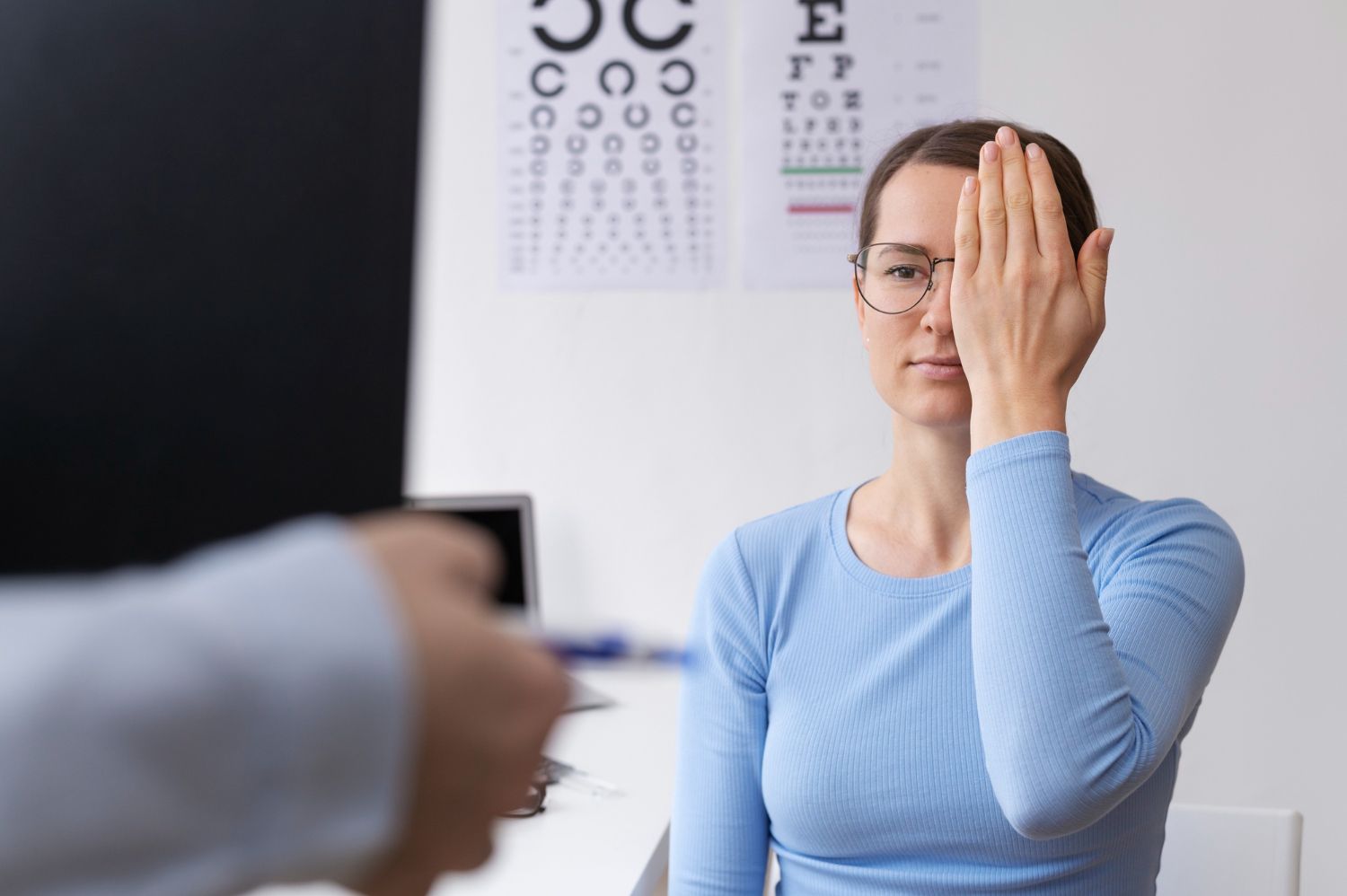 Woman in blue shirt covering one eye during an eye exam with an eye chart in the background