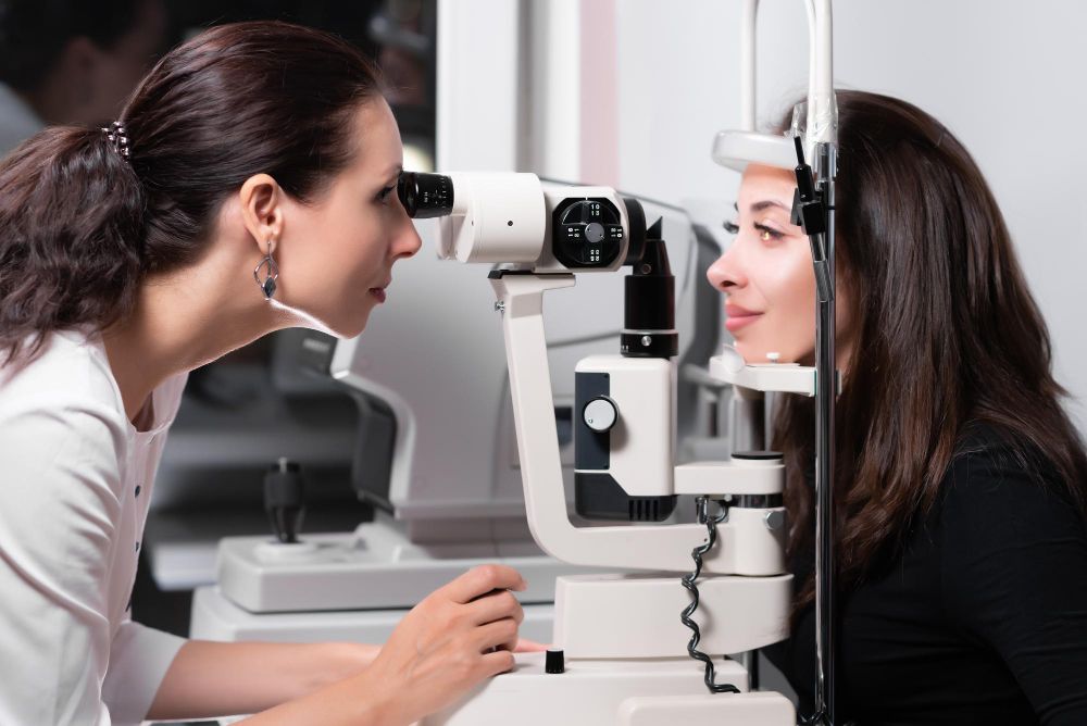 Two women using an eye exam machine in an optometry clinic, facing each other across the device