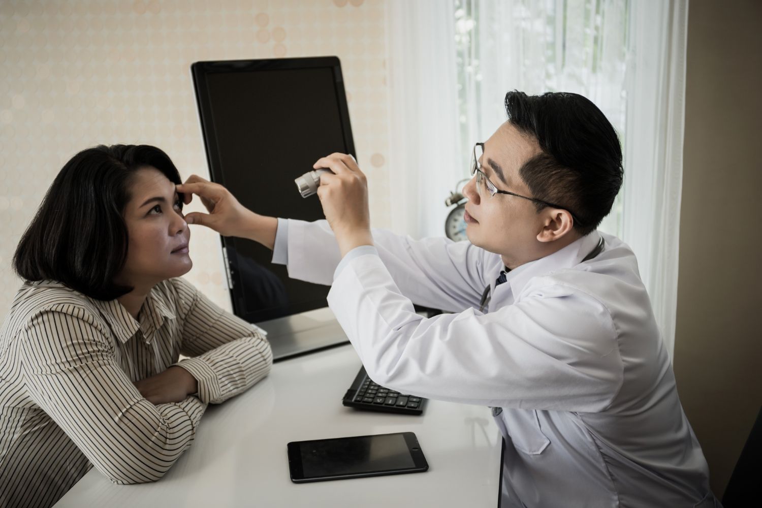 Eye doctor examining a patient’s eye in a clinic, with a chart and phone on the desk.