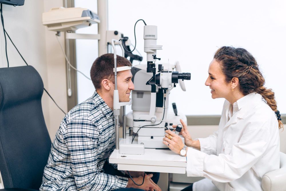 Patient and optometrist at an eye exam machine, facing each other and smiling in a clinic.