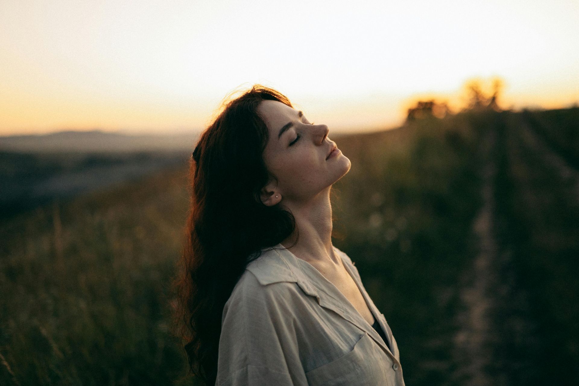 Woman in a beige shirt stands in a field at sunset, eyes closed and face turned toward the light.