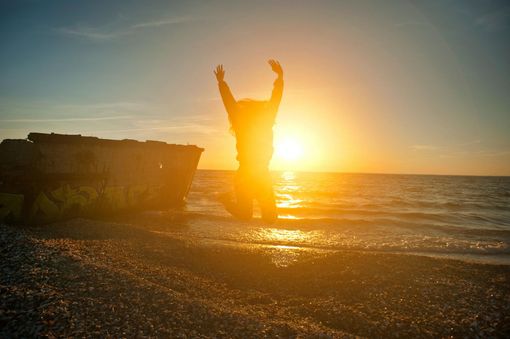 Silhouette of a person raising both arms at sunset on a beach with waves and a large rock nearby