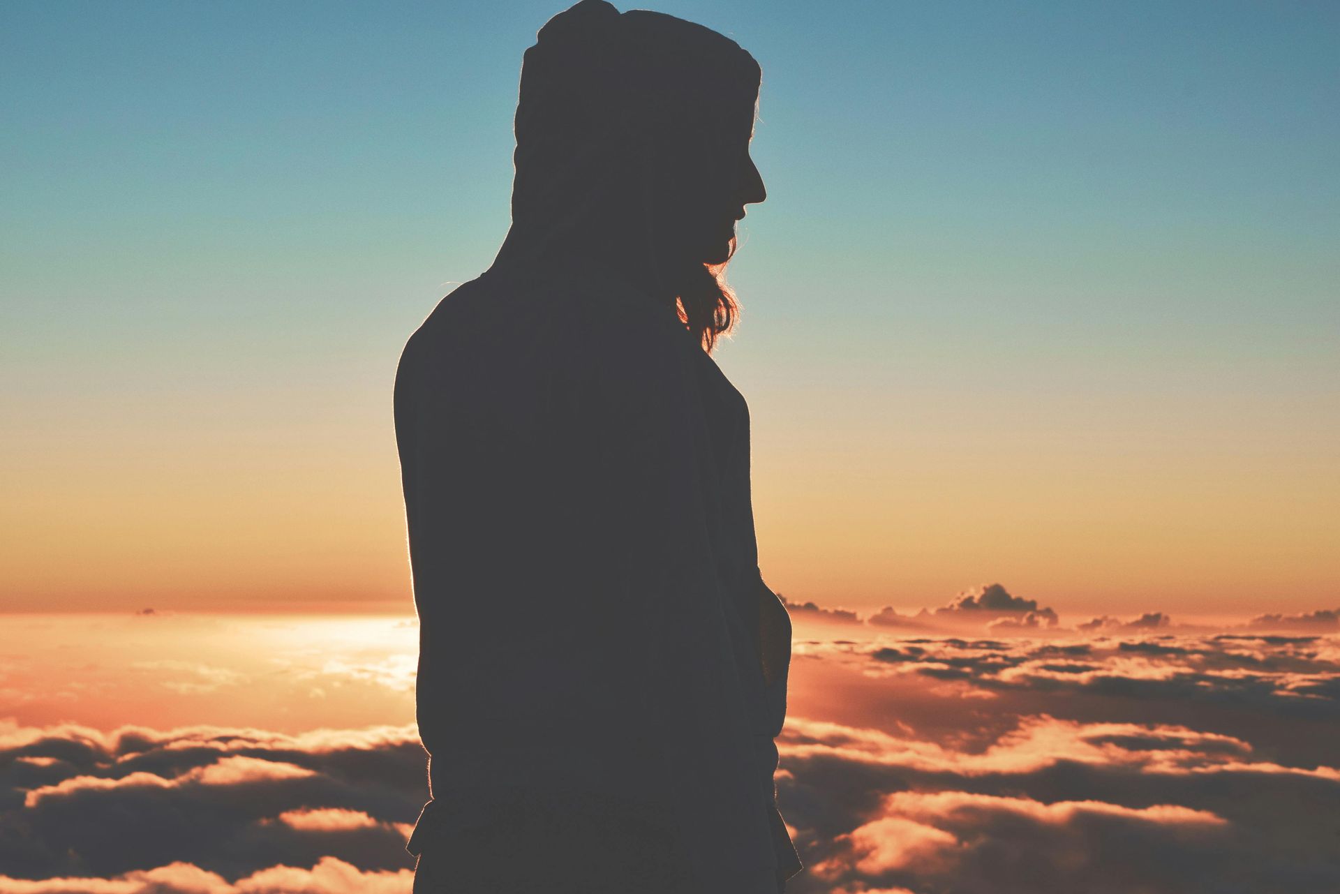 Silhouetted person standing above a sea of clouds at sunrise, with orange and blue sky trauma healing