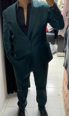 Persona con traje verde azulado oscuro y camisa negra tomándose una selfie frente a un espejo en el interior de un edificio.