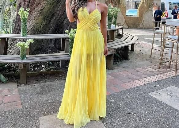 Mujer con un vestido amarillo brillante y vaporoso de pie al aire libre junto a un gran árbol y un banco de madera.