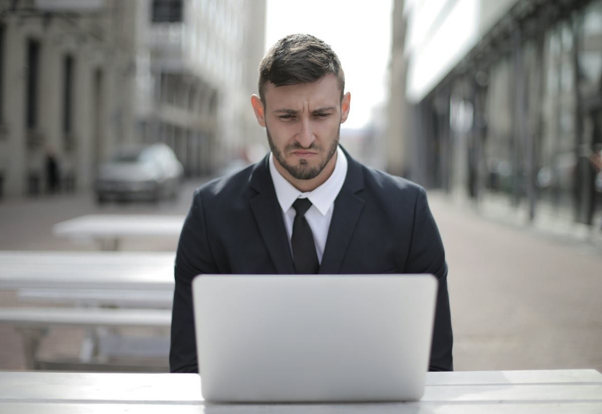 A person in a business suit with a furrowed expression works on a laptop at an outdoor table in a city setting.