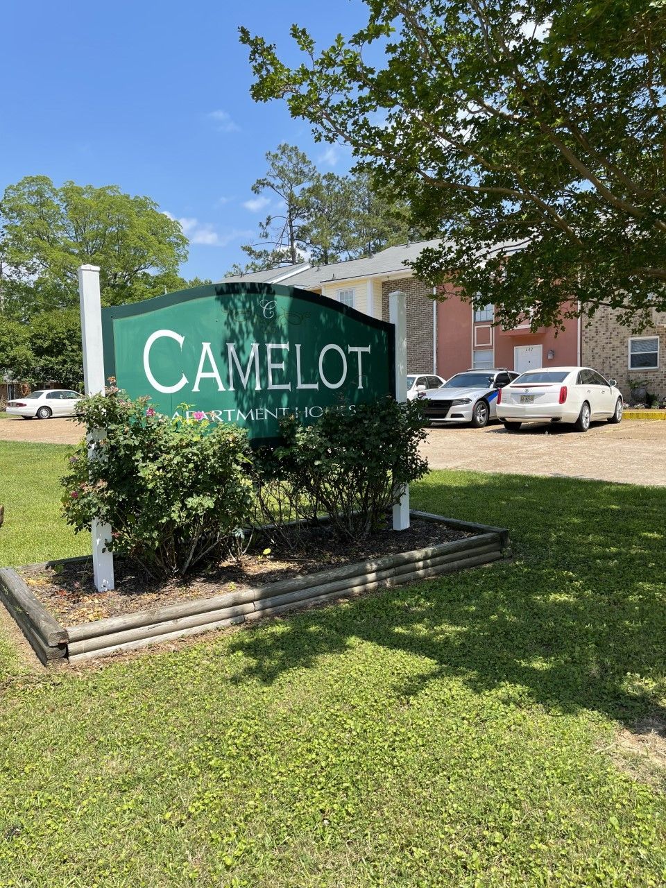 A green camelot sign is in the grass in front of a building.