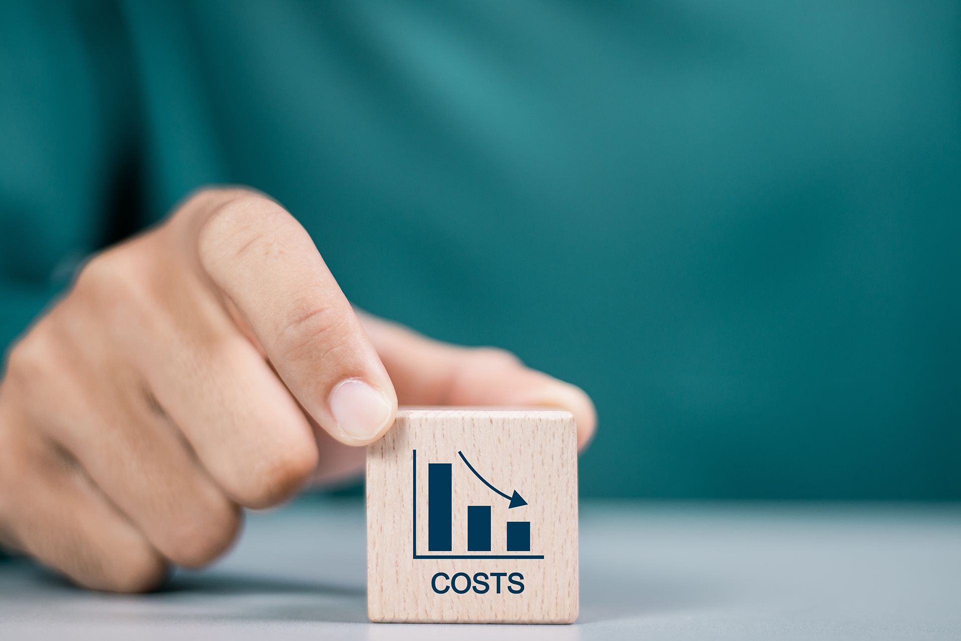 Hand holding a wooden block with a downward trending cost graph; turquoise background.