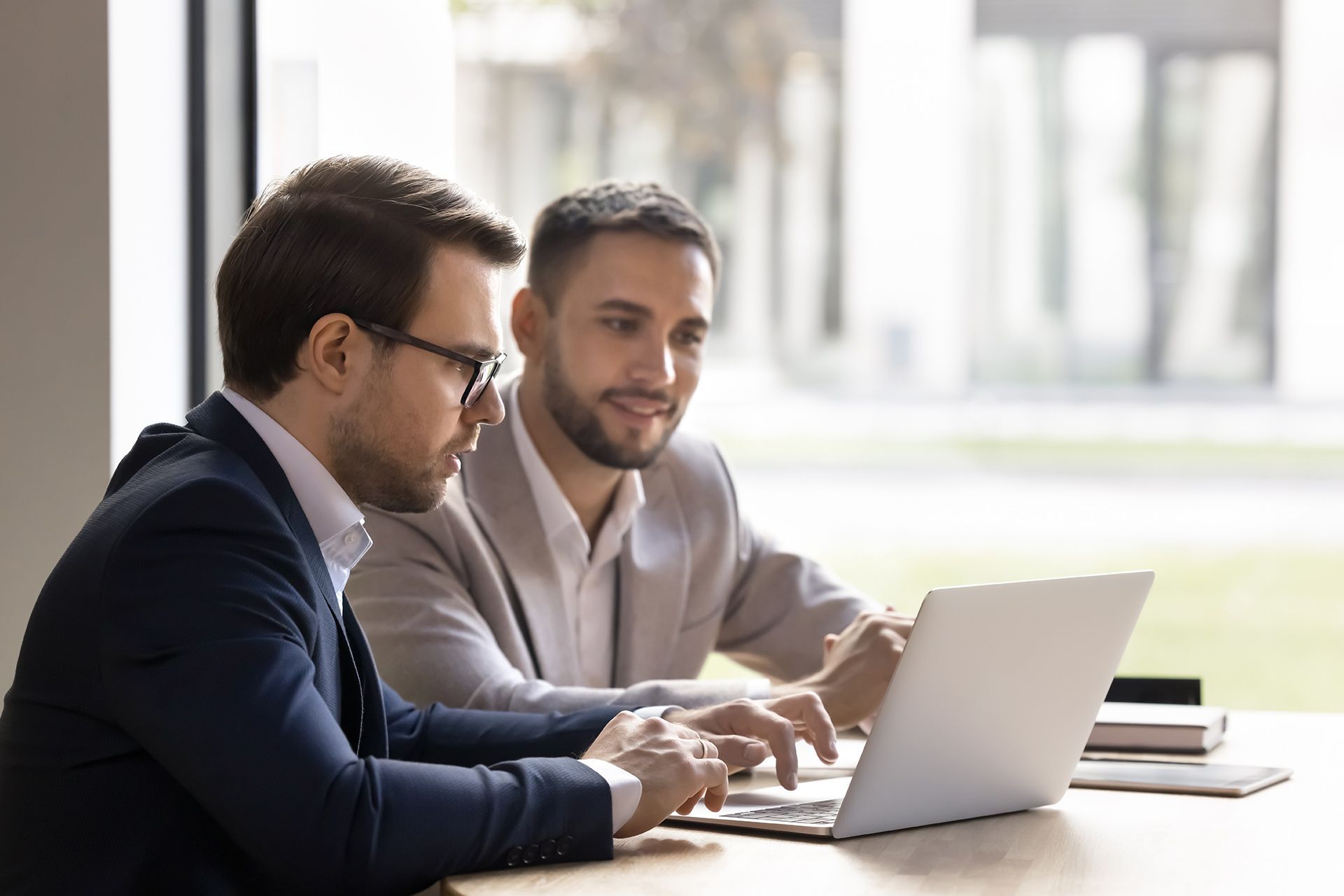 Two men in suits look at a laptop, one points, the other smiles.