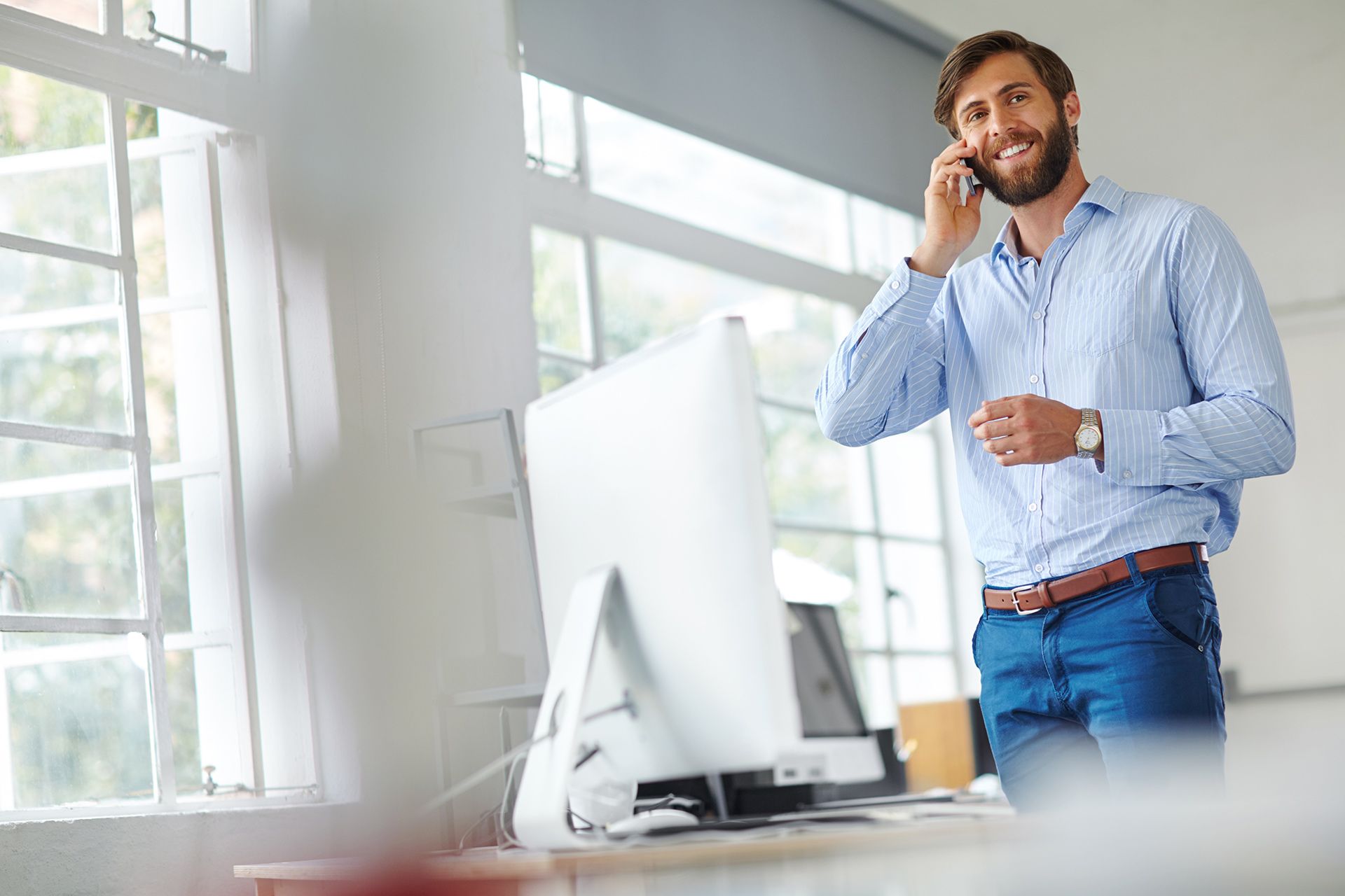 Man in light blue shirt and jeans, on phone, smiling near a window, office setting.
