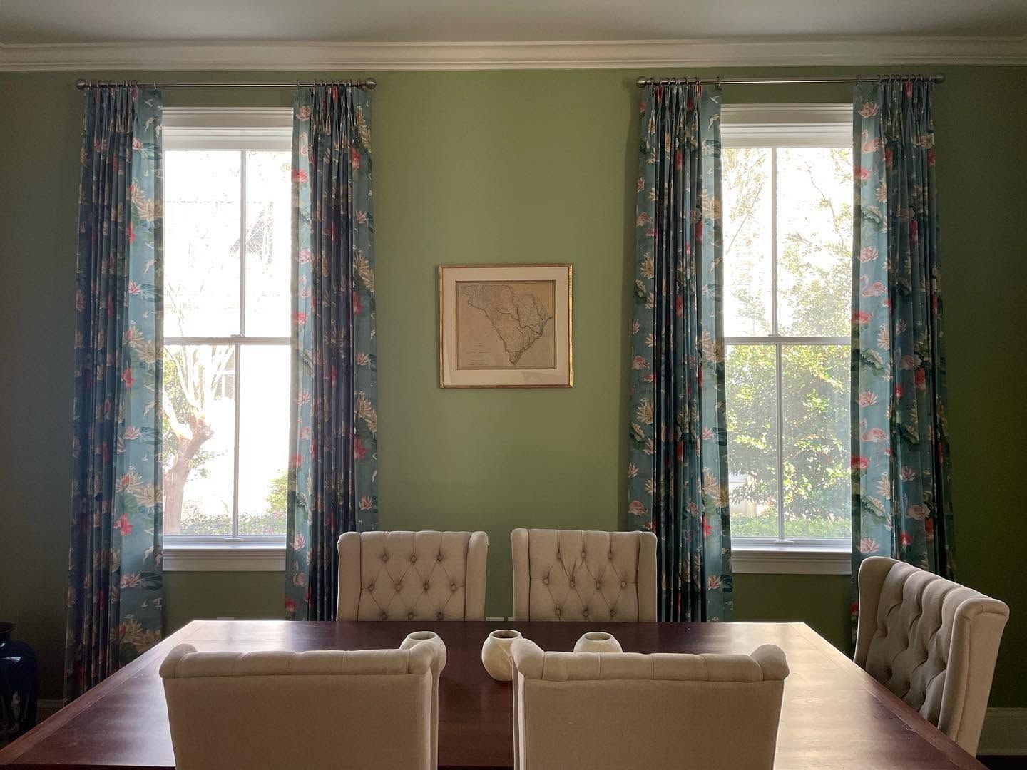Dining room with green walls, tall windows, patterned curtains, and a wooden table with beige chairs.