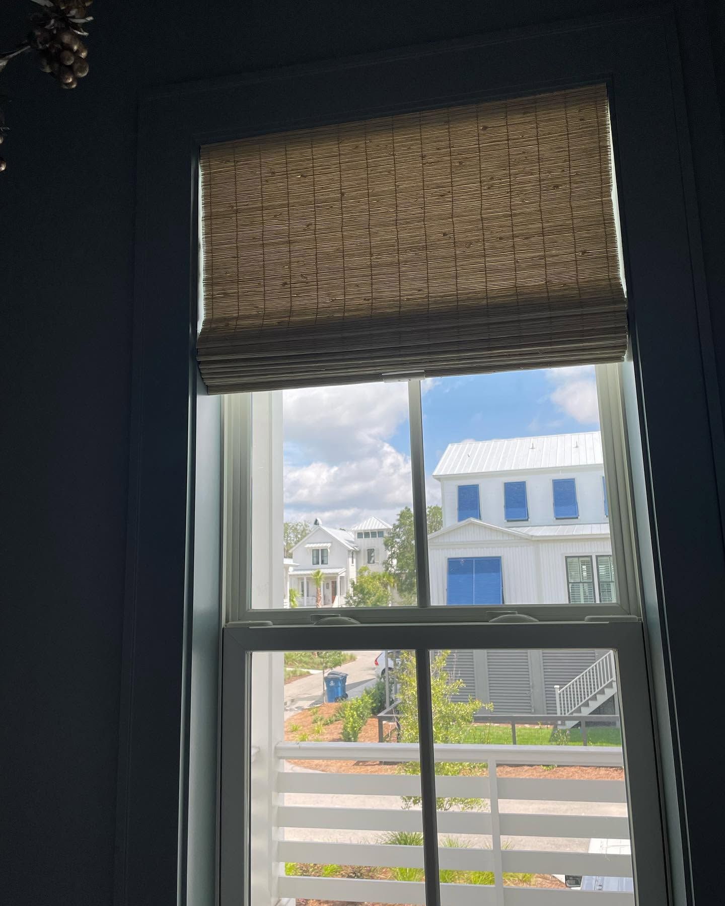 View through a window with a woven blind to a sunny street and white house with blue shutters.