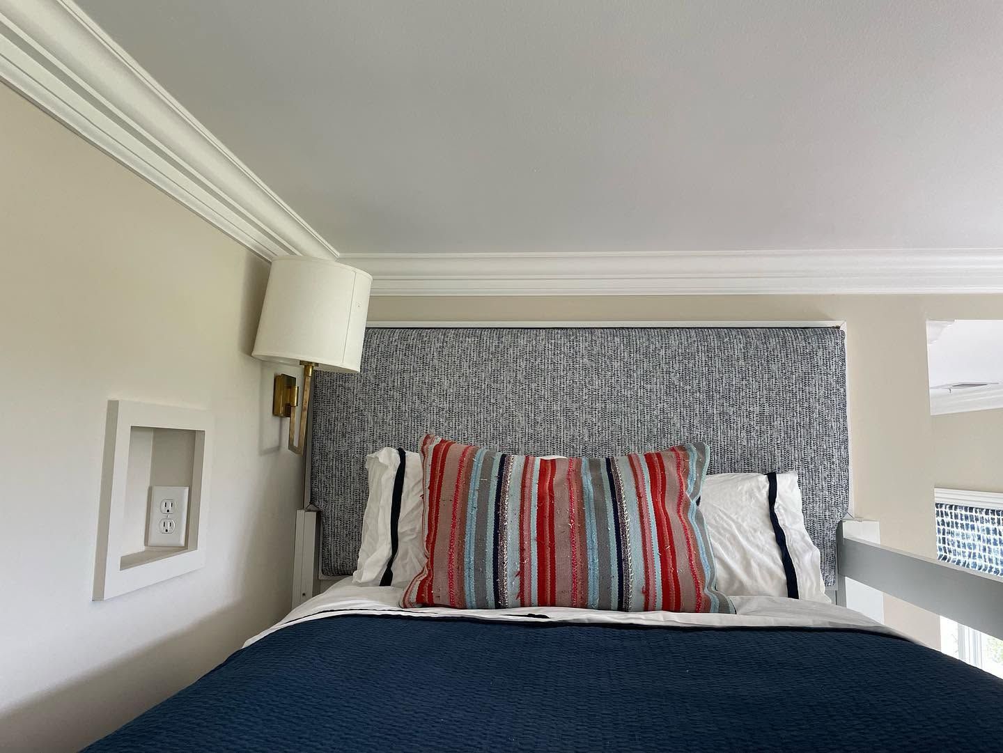 Bedroom corner with striped pillows, blue bedspread, wall lamp, and crown molding by a window