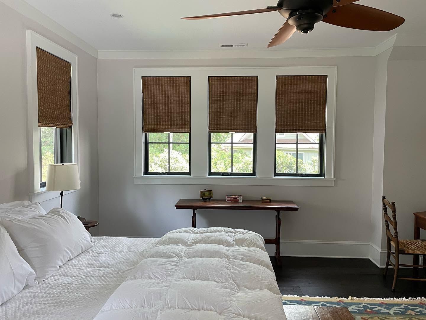 Bright bedroom with white bedding, bamboo shades, three windows, and a ceiling fan.