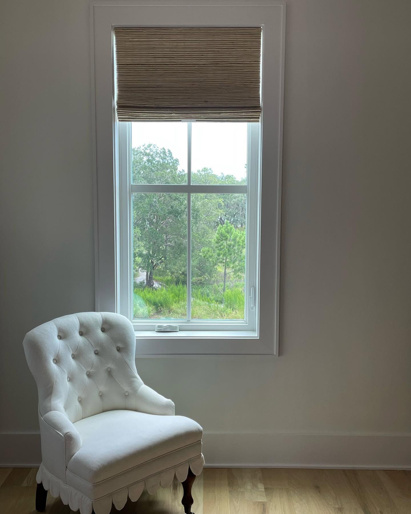 White tufted chair beside a tall window with a woven blind in a bright room.
